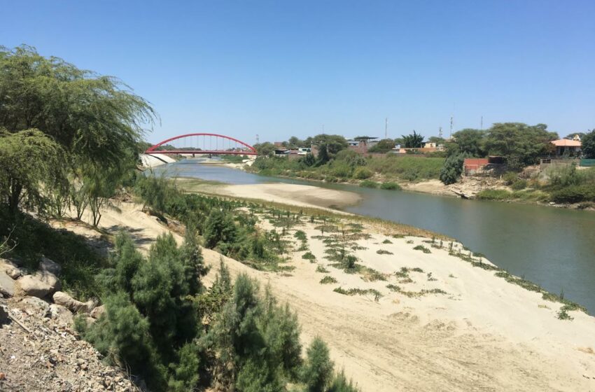View on the Piura River with green sidelines, blue sky and a red bridge in the background.