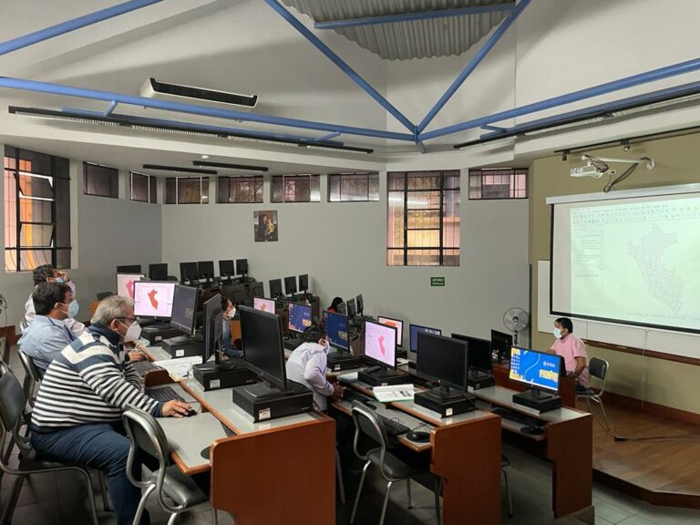 In a university room, several people sit in front of computers and listen to a man giving a training session in the front.