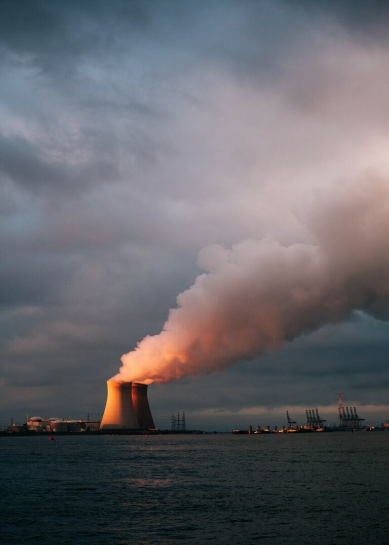 A nuclear energy plant at sunset exhausts big clouds of smoke into a cloudy grey sky.